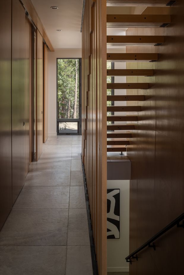 A refined residential hallway beside an open stairwell. The corridor has large pale stone or concrete-look floor tiles, warm timber wall panelling and vertical slatted screening, dark built-in cabinetry, recessed ceiling lights, and a full-height window overlooking trees. Open timber stair treads span the stair void, with dark handrails and a partial view of artwork below. The composition emphasizes warm natural materials, layered screening, and carefully controlled daylight.