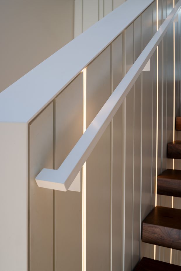 Close-up architectural photograph of a contemporary stairway. White-painted diagonal handrails and slim vertical balusters run alongside dark timber stair treads, with warm linear lighting integrated into a pale vertical-panelled wall.
