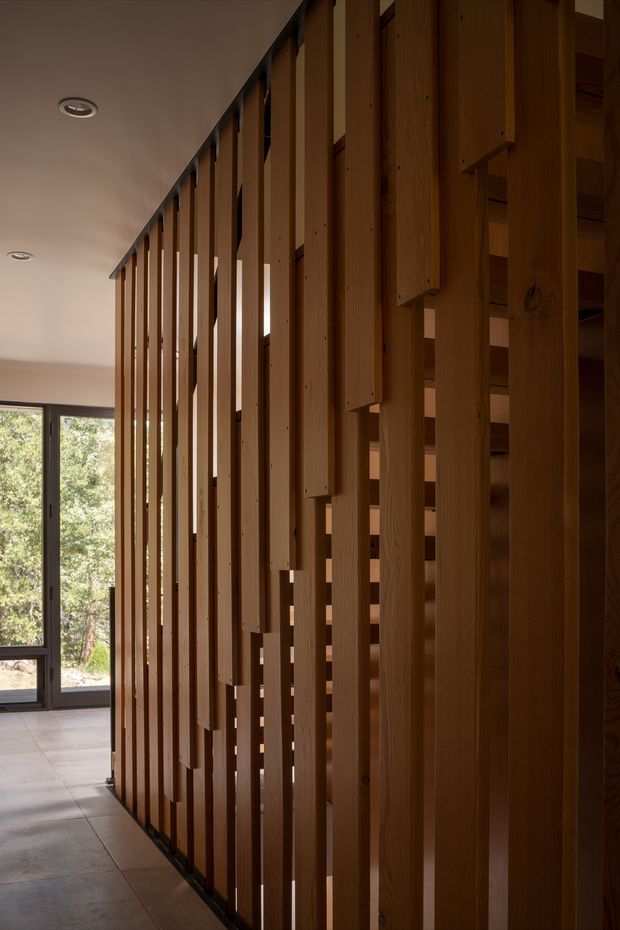 Interior architectural view of a warm timber slatted partition screening a staircase or circulation zone. Vertical timber battens create a rhythmic semi-transparent divider, with staggered solid timber elements visible behind. A large dark-framed glazed opening at the end of the space looks onto trees, while the floor is finished in large pale grey tiles and the ceiling has recessed downlights.