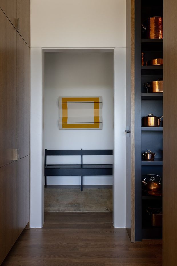A narrow, refined interior passage framed by full-height dark timber cabinetry. The central recessed alcove contains a built-in black bench with open shelving or drawers below, a concrete-toned floor area, and a contemporary geometric artwork. To the right, dark open shelving displays copper cookware and vessels. The space uses warm timber, white plaster, dark joinery, and metallic accents, creating a compact, gallery-like transitional zone.