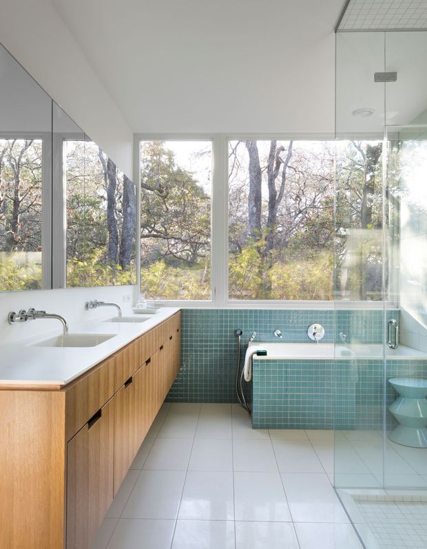 Bright modern bathroom with a long timber vanity and integrated white basins on the left, a built-in bathtub clad in small teal-green square tiles, and a glass shower enclosure on the right. Large windows overlook leafy trees, while pale floor tiles, white walls, chrome fixtures, and a mirrored wall create an airy, light-filled interior.