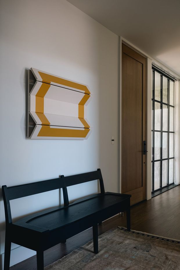 A refined residential entry hall with a black timber bench on a dark wood floor, a large geometric gold-and-white wall artwork, a tall timber door, and black steel-framed glazed doors bringing in natural light. The space has a restrained, contemporary character with strong geometric detailing and warm neutral finishes.
