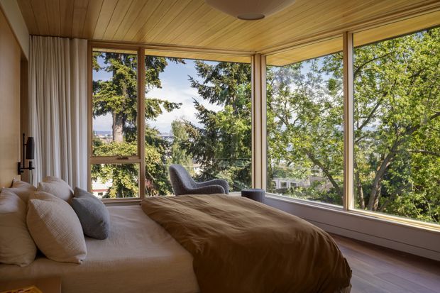 A warm, tree-filled bedroom with a low-profile bed, layered neutral bedding, a sculptural lounge chair, and expansive corner glazing overlooking leafy surroundings. The room features extensive timber panelling across the ceiling and a calm, nature-connected atmosphere.