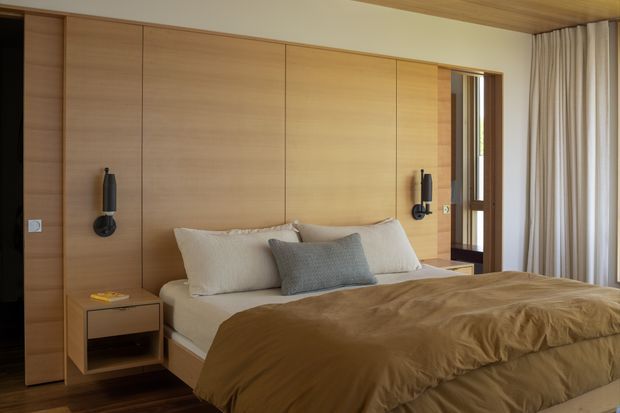 Warm contemporary bedroom with a large bed against a full-height timber-panelled feature wall. Built-in bedside niches and wall-mounted black reading lights flank the bed, while a timber ceiling, dark timber flooring, a glazed doorway, and full-height curtains complete the room.