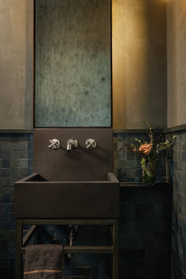 A moody, compact bathroom featuring a substantial dark stone or concrete trough basin on a dark metal frame, wall-mounted cross-handle tapware, deep blue-green square wall tiles, and textured warm grey plaster walls. A tall narrow mirror or reflective panel is mounted above the basin, with warm concealed lighting and a small floral arrangement adding contrast.
