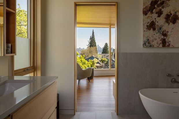 A warm, refined bathroom viewed toward an adjoining bedroom or sitting area through an open doorway. The foreground features a white vessel basin on light timber vanity cabinetry, a freestanding white bathtub, grey vertically textured wall tiles, and a floral artwork. Timber-framed glazing and a partially lowered timber blind frame a leafy suburban outlook, while the adjoining room has light timber flooring and a lounge chair.