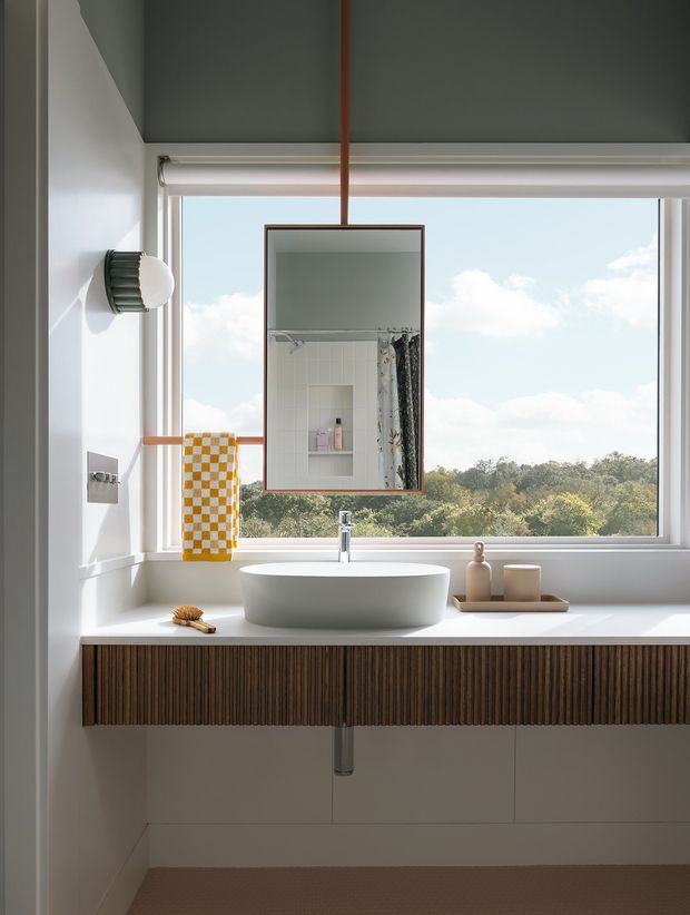 A bright, contemporary bathroom vanity positioned beneath a large picture window overlooking trees and sky. The space features a white vessel basin on a white countertop, a wall-mounted chrome tap, a vertically ribbed dark timber vanity front, a suspended rectangular mirror with a warm timber frame, white wall and floor finishes, and understated accessories. A shower area is visible in the mirror reflection.