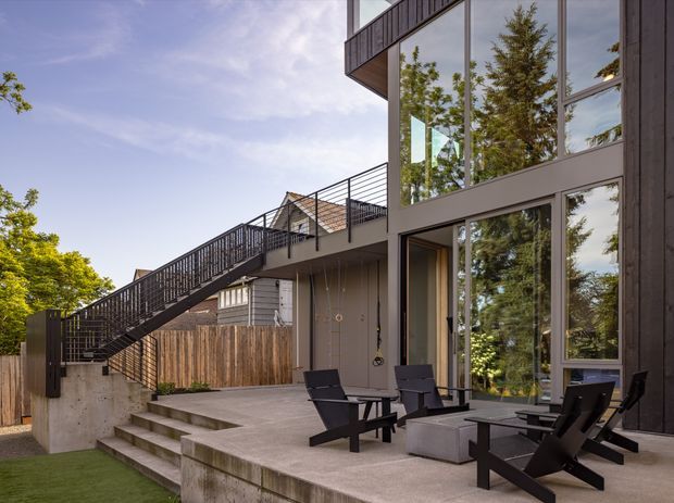 Exterior architectural photograph of a modern multi-level residential building with a large concrete terrace, broad floor-to-ceiling glazing, dark vertical cladding, exposed concrete steps and retaining walls, and a prominent black metal exterior stair and balcony railing. Outdoor lounge chairs and a low table occupy the terrace, with mature trees reflected in the windows and visible around the building.