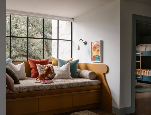A warm, thoughtfully styled bedroom reading nook with a built-in timber window seat and upholstered cushion beneath large black-framed grid windows. The seat includes integrated storage cabinetry and curved timber end panels, with layered neutral, rust, teal and mustard cushions, a plush toy, wall-mounted sconce and colourful artwork. A doorway at right reveals a secondary room with bunk beds. The space has white walls, dark timber flooring and a cozy, mid-century-influenced character.