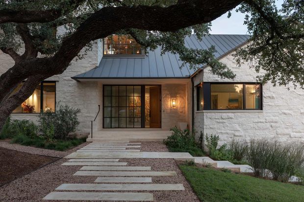 Exterior view of a low-rise contemporary home set beneath a mature spreading tree. The facade combines pale textured masonry walls, dark standing-seam metal gabled roof sections, large black-framed windows, and a timber entry door. A layered concrete stepping-stone path crosses gravel and planted garden beds toward the recessed front entrance, with warm interior lighting visible through the glazing.