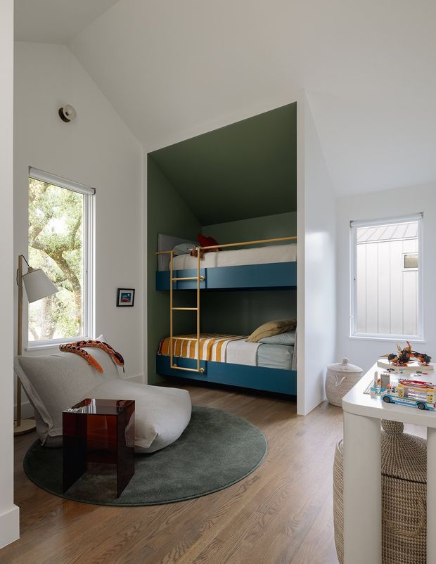 A bright, compact children's bedroom with a dedicated recessed bunk-bed alcove. The alcove is painted deep green and fitted with two blue-fronted bunk beds and a yellow ladder/guard rail. The main room has white walls, light timber flooring, a sculptural pale lounge chair, a dark green oval rug, a small dark side table, and a white children's desk with woven storage baskets and toys. Large windows provide natural light and views of trees and a neighbouring structure.