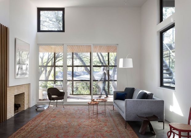 Bright double-height living room with expansive floor-to-ceiling glazing, a brick fireplace, dark timber flooring, a large patterned area rug, and contemporary seating. Tall windows overlook leafy surroundings, while a sculptural floor lamp and restrained furnishings create an airy, refined interior.