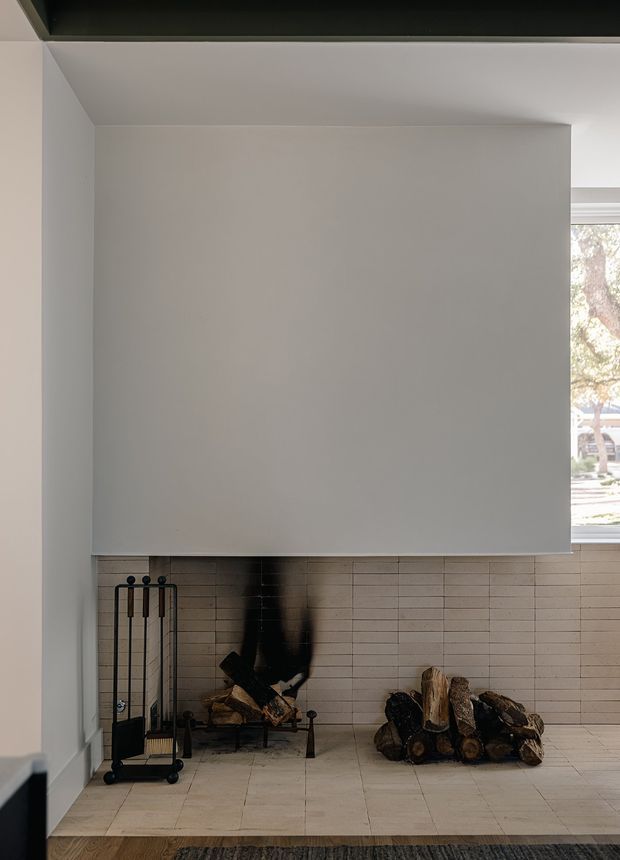 Minimalist interior view of a built-in fireplace alcove. A large smooth painted-plasterboard rectangular surround projects over a light brick-tile fireplace wall, with a dark firebox, stacked firewood, and black metal fire tools. Pale tiled flooring and walls create a restrained neutral scheme, while a window at right brings in daylight and a glimpse of the exterior.