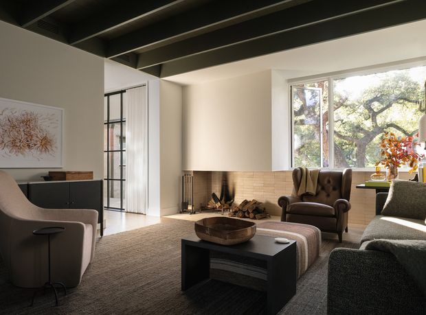 Warm, refined living room with a large sectional sofa, upholstered lounge chair, tufted leather armchair, central coffee table, and a modern open fireplace clad in light brick. Full-height glazing and a black-framed glazed door bring in abundant natural light, while dark exposed ceiling beams create strong architectural contrast.