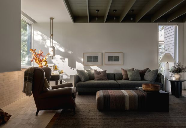 A warm, refined living room with a large dark sectional sofa, leather lounge chair, striped upholstered ottoman, side tables, framed artwork, and a floor lamp. Strong sunlight enters through large windows, highlighting pale walls and a feature ceiling with exposed dark beams and recessed lighting. A low light-brick surface appears along the left side, while greenery is visible outside.
