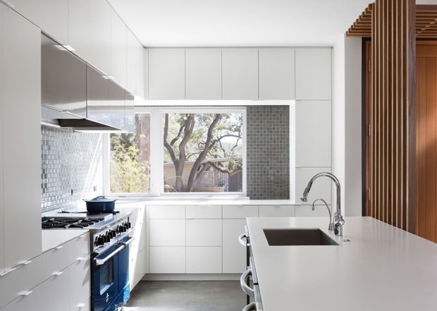 A bright, contemporary white kitchen with extensive handleless cabinetry, a large island with integrated dark sink and sculptural chrome tapware, and a freestanding blue range cooker. The kitchen features a grey mosaic-tile splashback around the window, grey concrete-look flooring, and a striking vertical timber screen or battened partition at the right. Natural light enters through the large window, creating a clean, refined interior.