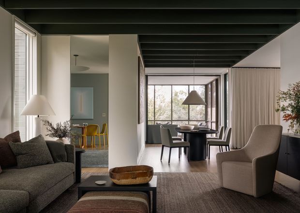 A refined open-plan interior combining a living room and dining area. The space features a dark exposed-beam ceiling, pale plaster walls, large windows, a textured grey sofa, sculptural lounge chair, dark dining table and chairs, and restrained contemporary furnishings. A small kitchen or kitchenette area with yellow stools is visible beyond the living space.
