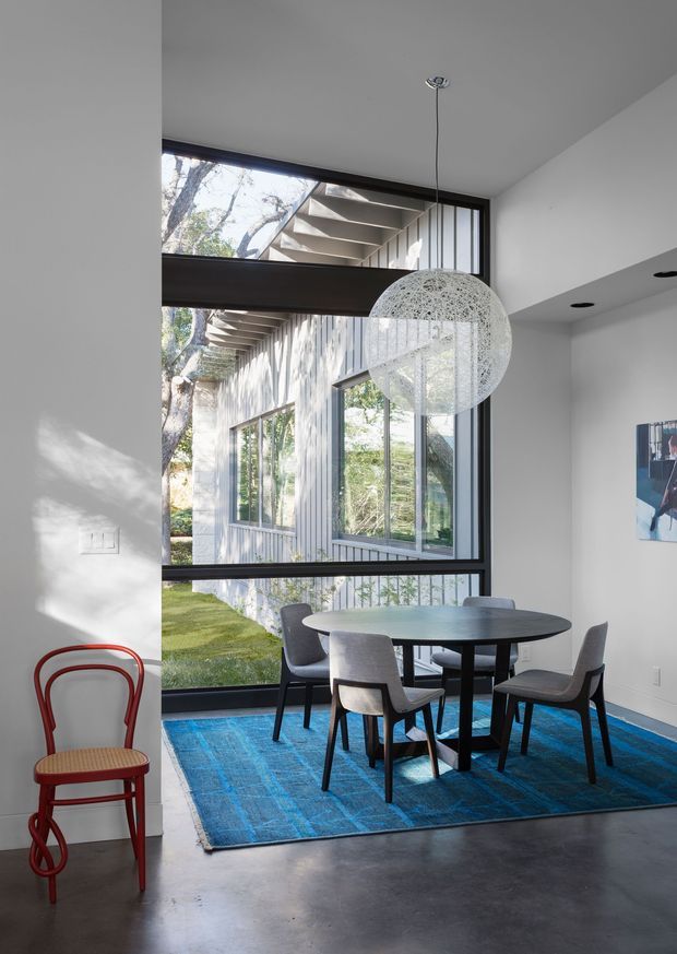 A contemporary dining area with a round dark dining table and four upholstered chairs positioned beside expansive black-framed glazing. The room features a polished concrete floor, a vivid blue area rug, white walls, and a large spherical pendant light. Through the glazing, a landscaped courtyard and the building's textured exterior with projecting roof fins are visible. A red bentwood-style chair adds a bright accent at the side.