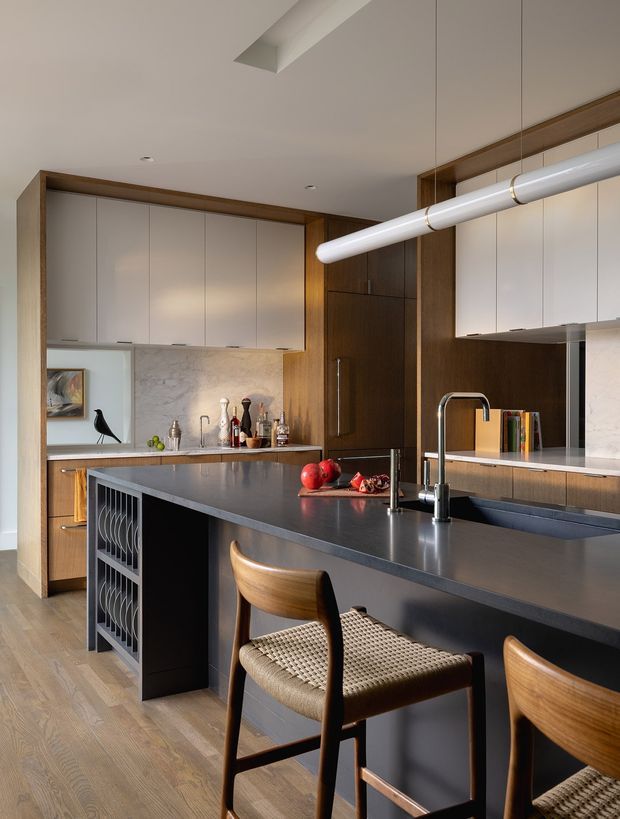 A refined contemporary kitchen with a large dark island and integrated sink, warm timber cabinetry and framing, pale flat-panel overhead cupboards, and a light stone or marble-look backsplash. The island includes open wine or bottle storage and seating for several people on woven-seat timber stools. A linear white pendant light runs above the island, while timber flooring and carefully integrated appliances complete the sophisticated interior.