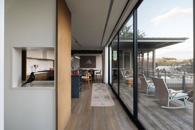 Contemporary open-plan interior with a long timber-finished floor and full-height black-framed glazing opening onto a covered timber terrace. The space includes a compact kitchen and dining area, a timber feature wall with recessed display niche, and outdoor lounge seating visible through the glazing. The design emphasizes indoor-outdoor connection, clean linear detailing, and natural materials.