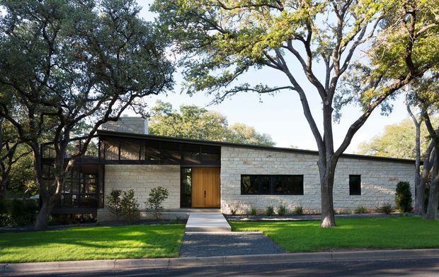 Single-storey modern residence viewed from the street, set among mature trees and a neatly landscaped lawn. The building has a low horizontal profile, pale coursed masonry walls, dark-framed glazing, a broad glazed clerestory-like section, and a warm timber entry door. A simple paved path leads from the sidewalk to the entrance.