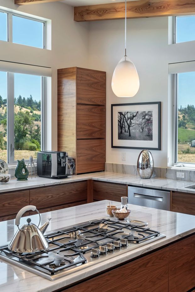 A bright, contemporary kitchen with a large central island featuring an integrated gas cooktop and pale marble benchtop. Rich walnut cabinetry, a tall walnut storage unit, white walls, exposed timber ceiling beams, expansive windows, and a sculptural pendant light create a warm, refined interior. A rural, tree-covered landscape is visible through the windows.