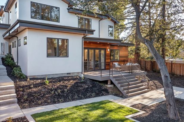 Exterior view of a contemporary two-storey home with white rendered walls, dark window frames, projecting roof eaves, and a warm timber-clad recessed entry or outdoor living area. A raised timber deck with metal balustrades and stairs connects to concrete paths, while mature trees and a timber boundary fence surround the landscaped yard.