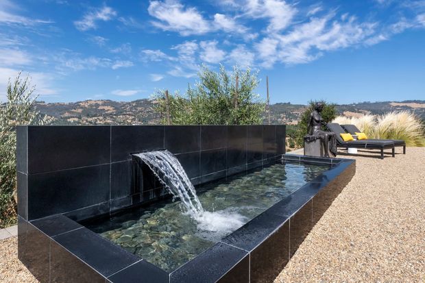 A contemporary outdoor courtyard or terrace featuring a long rectangular reflecting pool with a sculptural water cascade, clad in dark glossy stone or tile. Gravel paving, ornamental grasses, lounge chairs, and a mountainous landscape surround the pool under a bright blue sky.