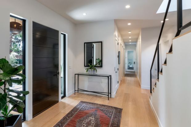 A bright, contemporary residential entry hall viewed from inside. The space features a dark timber front door with sidelights, pale timber flooring, white walls, a slim black-framed console table beneath a rectangular mirror, recessed ceiling lights, indoor greenery, and a staircase with a black balustrade leading upward. A patterned rug adds muted red and blue tones in the foreground, while a hallway extends toward a bathroom or utility area.