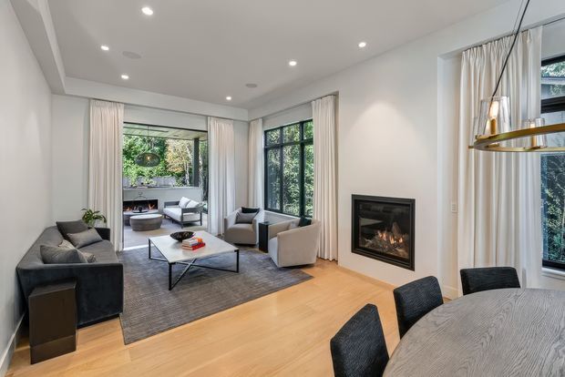 Bright, refined open-plan living and dining room with pale timber flooring, white walls, tall windows dressed in sheer curtains, a contemporary fireplace, grey upholstered seating, and a large round dining table. A secondary sitting area is visible through the wide opening at the rear, with abundant greenery outside the glazing.