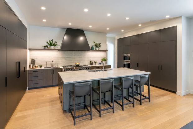 A spacious contemporary kitchen with extensive dark flat-panel cabinetry, a large central island with seating for four, integrated appliances, a stainless-steel range and oversized dark rangehood. Pale timber flooring, white walls and a light stone island benchtop create a bright, refined contrast with the dark joinery. Recessed ceiling lighting and indoor plants add to the clean, modern fit-out.