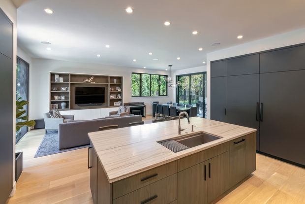 Bright open-plan contemporary kitchen and living area. A large timber island with integrated sink and dark fixtures dominates the foreground, with extensive dark built-in cabinetry along the right wall. The adjoining living area includes a sofa, built-in media shelving and television, while a dining table is visible near glazed doors opening to greenery outside. Recessed ceiling lights, pale walls and timber flooring create a clean, warm interior.