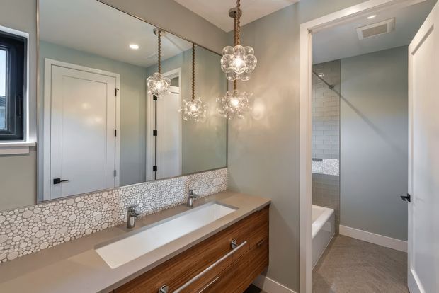 A refined bathroom vanity area with a long wood-grain floating cabinet, pale stone countertop, integrated white rectangular basin, and large wall-to-wall mirror. A patterned pebble mosaic backsplash runs beneath the mirror, while clustered glass pendant lights hang from the ceiling. A separate tiled shower and bathtub area is visible through the open doorway.