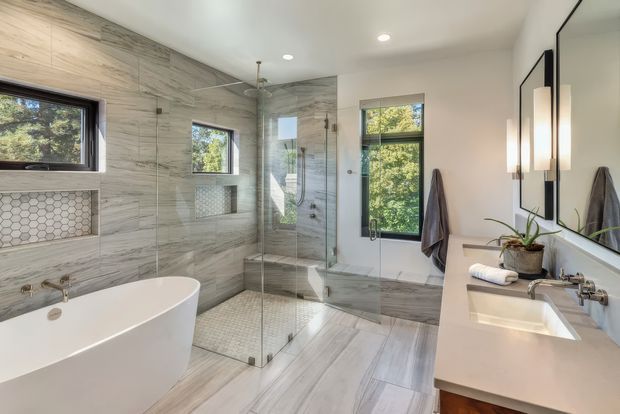 Bright, spacious contemporary bathroom featuring a freestanding white bathtub, glass-enclosed walk-in shower, wall-mounted toilet, double vanity, large mirror, and extensive grey marble-look tile finishes. Black-framed windows bring in views of greenery, while recessed ceiling lights and vertical wall sconces provide clean modern illumination.