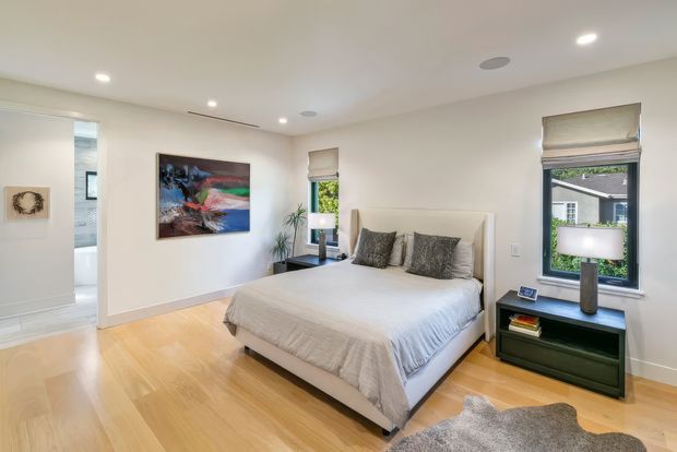 Bright, contemporary bedroom with a large upholstered bed, neutral bedding, dark patterned cushions, and a dark green bedside table. The room features pale timber flooring, white walls and ceiling, recessed lighting, roller blinds, artwork, and windows overlooking neighbouring buildings and greenery. An open doorway leads toward a tiled bathroom or hallway.