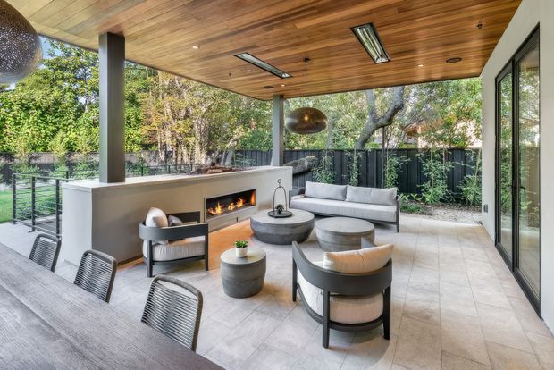 A refined covered outdoor living and dining area with a warm timber-lined ceiling, large glazed openings, tiled flooring, and a built-in linear fireplace. The space includes a dining table and chairs in the foreground, a lounge arrangement with curved upholstered chairs and sofas, a round coffee table, sculptural pendant lighting, and views to a landscaped garden beyond. The design blends indoor comfort with an open-air pavilion feel.