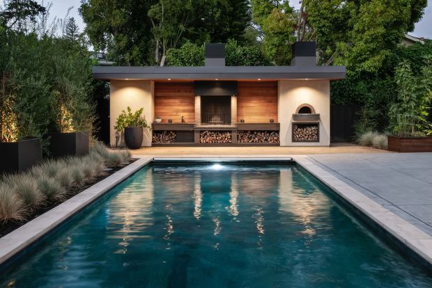 A landscaped outdoor pool area with a long rectangular swimming pool in the foreground and a covered outdoor entertaining pavilion at the far end. The pavilion features a central built-in fireplace or grill, a wood-lined niche, stacked firewood storage, a secondary arched fireplace, dark flat roof forms, and warm integrated lighting. Large concrete pavers, ornamental grasses, sculptural planters, and dense greenery frame the space.