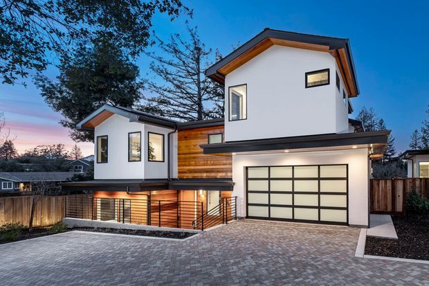 Contemporary multi-level residence photographed at dusk, featuring crisp white rendered walls, dark roof and trim, warm timber cladding, large black-framed windows, a projecting garage, and a broad grey brick-paved driveway. The split-level form and elevated lower level are clearly visible, with a fenced suburban setting and landscaped edges.