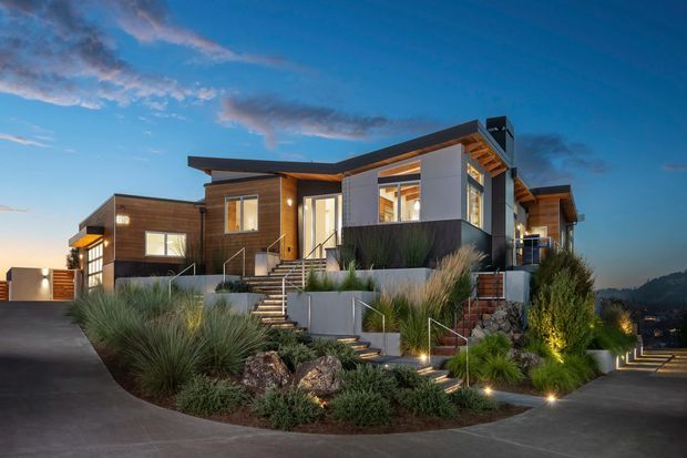 A contemporary multi-level hillside residence photographed at dusk. The home has a strong horizontal composition with offset flat and low-pitched roof planes, dark timber-clad walls, light rendered wall sections, extensive glazing, and exposed timber soffit details. Broad concrete steps and retaining walls connect the house to a landscaped driveway, with ornamental grasses, shrubs, rocks, and integrated low-level lighting creating a dramatic approach.