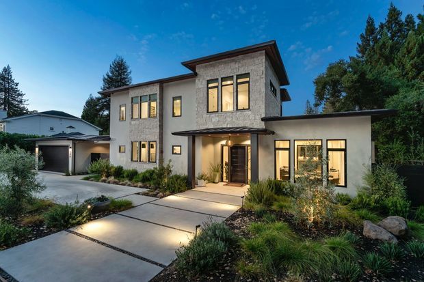 Contemporary two-storey residence viewed from the landscaped front approach at dusk. The home has a flat-to-low-pitched roof composition with broad eaves, pale stucco walls, a prominent textured stone-clad upper facade, dark-framed windows, and a recessed timber front door. A connected carport/garage structure sits to the left, while concrete stepping slabs and low, drought-tolerant planting define the entry garden.