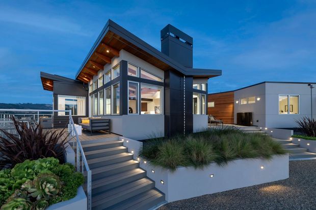 Exterior dusk view of a contemporary multi-level residence with a steep asymmetrical roof, extensive floor-to-ceiling glazing, dark metal cladding, timber soffits and wall panels, and a prominent chimney-like vertical element. Illuminated timber-and-metal stairs lead through landscaped raised planters to the house, with a deck and distant waterfront or landscape visible beyond.