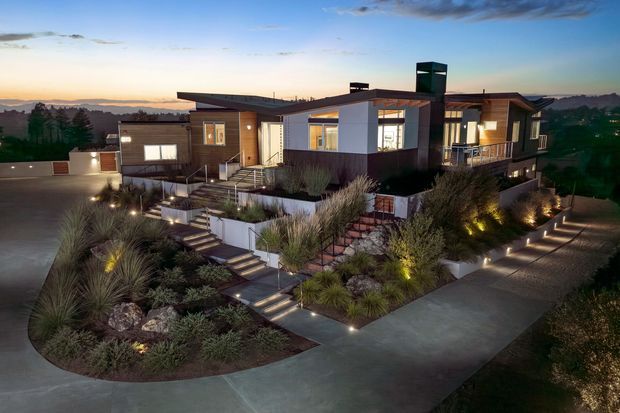 Twilight exterior view of a contemporary single-storey home with a low-pitched roof, varied timber and light-coloured wall cladding, extensive glazing, and a prominent dark chimney or vertical feature. The house is set within a carefully designed landscaped garden featuring ornamental grasses, rocks, low planting, concrete paths, steps, retaining walls, and integrated low-level lighting.