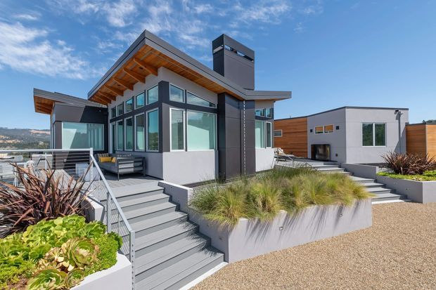 Exterior view of a contemporary multi-level residential home on a landscaped, sloped site. The house features extensive dark-framed glazing, projecting flat roof planes with exposed timber soffits and rafters, a tall dark chimney or fireplace volume, light rendered walls, timber cladding, raised concrete planters, and grey deck stairs. Drought-tolerant ornamental grasses and succulents surround the entry and terraces.
