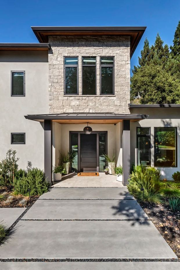 Front exterior view of a contemporary two-storey house. The facade combines pale rendered walls with a prominent light stacked-stone-clad upper volume, dark-framed windows, deep roof overhangs with timber soffits, and a sheltered entry porch with a dark timber door and pendant light. A broad concrete path with gravel joints leads through minimalist drought-tolerant landscaping and ornamental grasses to the entrance.