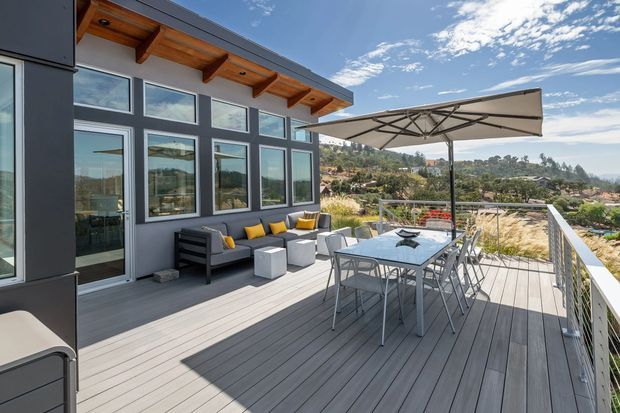 A spacious elevated outdoor deck adjoining a modern home, with grey composite-style decking, dark-framed floor-to-ceiling windows, timber-lined exposed eaves, and a large retractable shade canopy. The deck contains an outdoor dining table with metal chairs and a lounge setting with dark seating and yellow accent cushions. A suburban hillside landscape is visible beyond the balustrade under a bright blue sky.