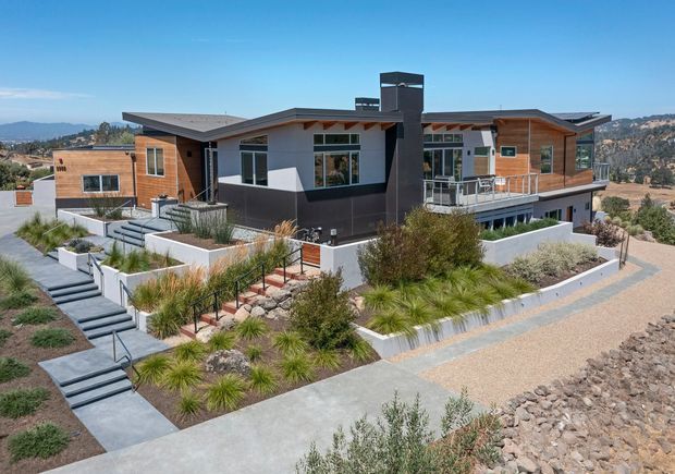 A contemporary multi-level residential house viewed from the landscaped front approach. The home features a low-pitched roof with broad overhangs, a mix of dark and light rendered walls with warm timber cladding, expansive windows, a prominent dark chimney or vertical feature, and balcony railings. Terraced concrete paths, stepped access, gravel paving, retaining walls, and drought-tolerant ornamental planting surround the house. A dry, hilly landscape is visible in the background.