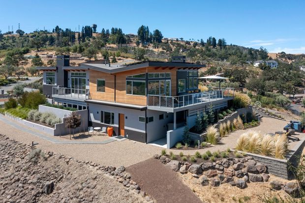 Exterior view of a contemporary two-storey house on a dry, gently sloping site. The home has a low-pitched roof with broad overhangs, warm timber cladding on the upper level, dark painted lower-level walls, expansive glazing, and a large elevated terrace with metal balustrading. Gravel paths, stone retaining elements, ornamental grasses, and drought-tolerant landscaping surround the building, with a hilly semi-rural landscape beyond.