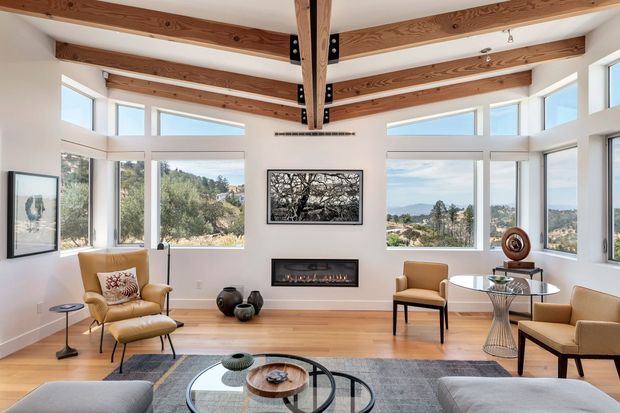 Bright contemporary living room with expansive corner windows, panoramic hillside views, exposed timber ceiling beams with dark metal connectors, a linear built-in fireplace, light timber flooring, and neutral modern furnishings arranged around a glass coffee table.
