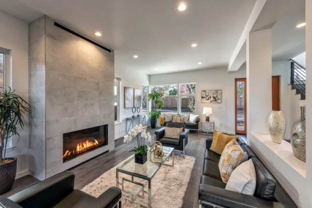 Bright, contemporary open-plan living room with a large tiled fireplace and built-in firebox as the focal point. The room includes black leather sofas and armchairs, a glass coffee table, a pale textured rug, indoor plants, artwork, recessed ceiling lights, and a decorative display niche. Large windows bring in natural light, while a stairway is visible at the rear.