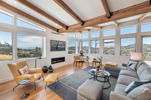 Bright, spacious living room with expansive corner glazing and scenic hillside views. The room features exposed timber ceiling beams with black metal connectors, pale timber flooring, a large light-grey sectional sofa, tan leather lounge chair and ottoman, layered round coffee tables, a grey area rug, a slim linear fireplace, and contemporary artwork. The interior has a warm, refined, mid-century-influenced feel with strong indoor-outdoor visual connection.